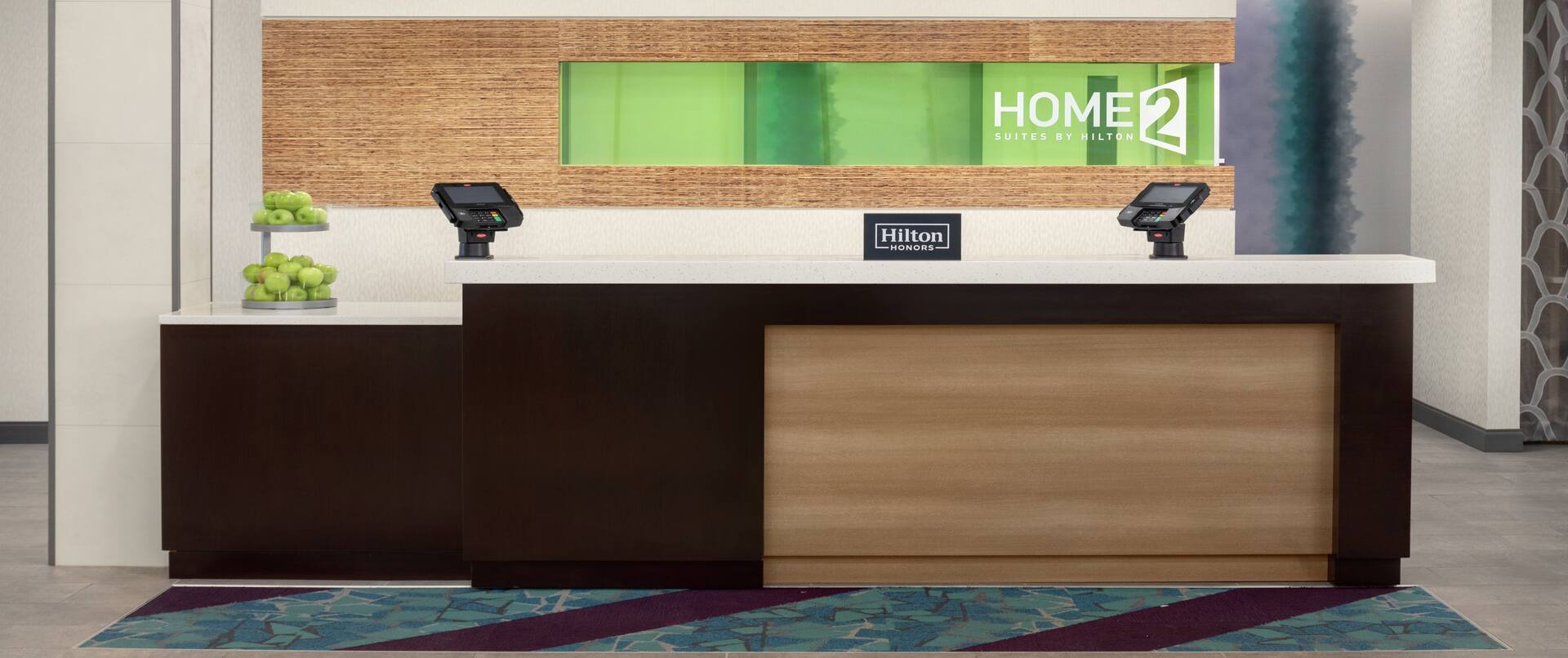 Welcoming front desk in hotel lobby featuring hilton honors sign, lowered accessible desk, and green apples.