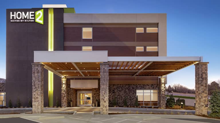 Modern Home2 Suites hotel exterior featuring porte cochere, glowing lobby, and beautiful dusk sky.