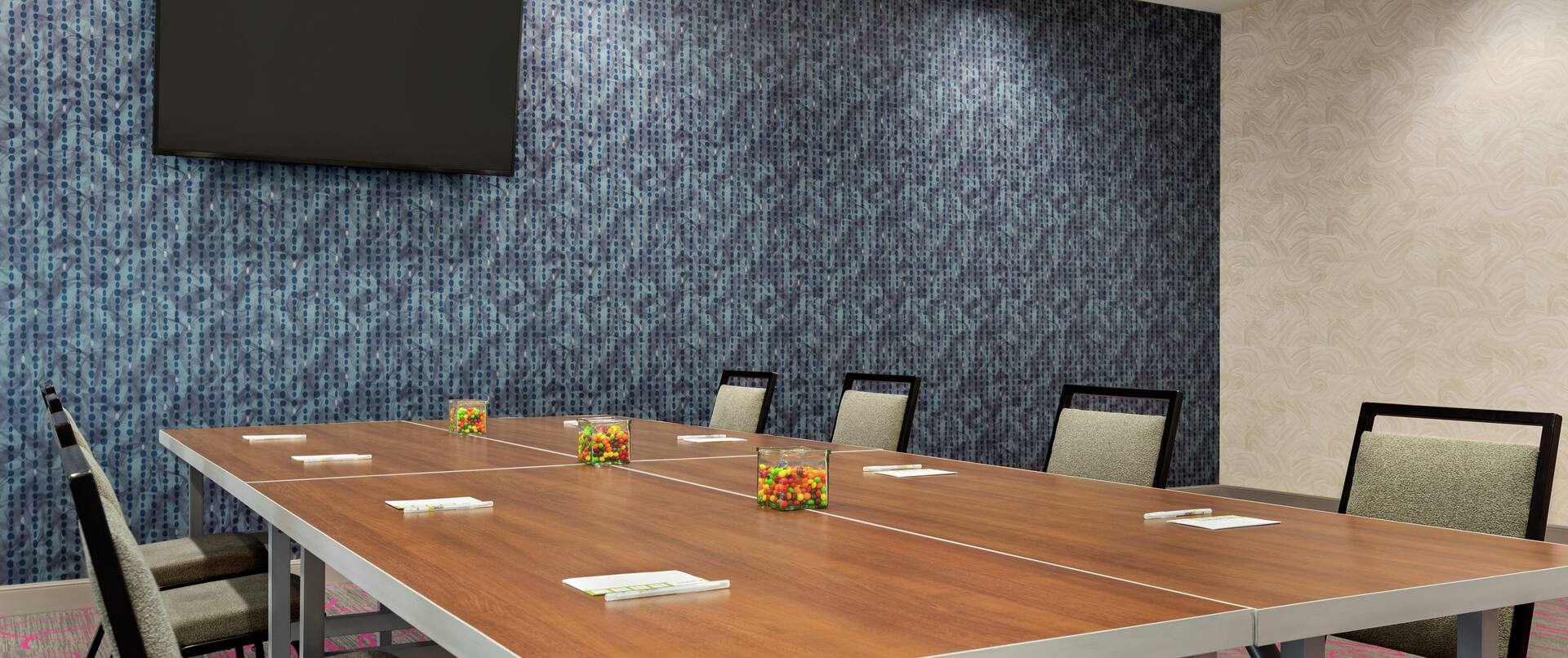 Spacious meeting room with boardroom table, notepads and pens, and TV on wall.