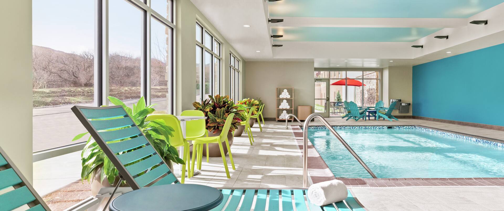 Spacious indoor swimming pool featuring floor to ceiling windows with beautiful mountain view, chaise lounge chairs, lush plants, and complimentary towels.
