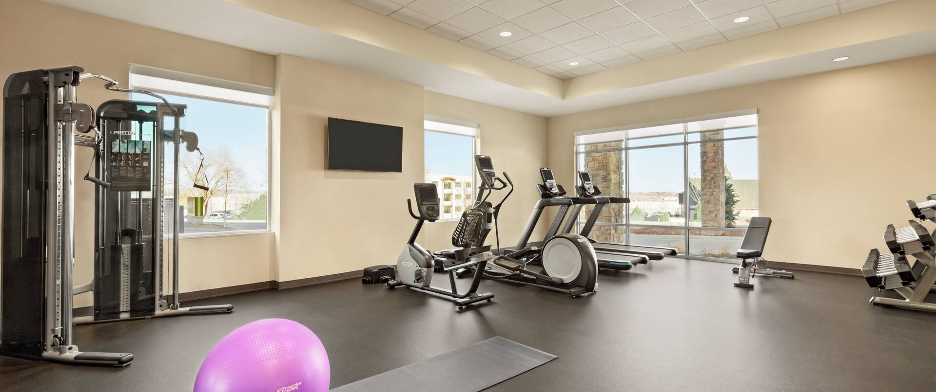 Spacious fitness center featuring floor to ceiling windows, cardio machines, free weights, yoga mats, and TV.