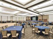 Wilson Hall Banquet Set Up With Dance Floor Surrounded by Mixed Tables With Blue Linens, Tan Chairs, and Large Presentation Screen With Black Drapes in the Background