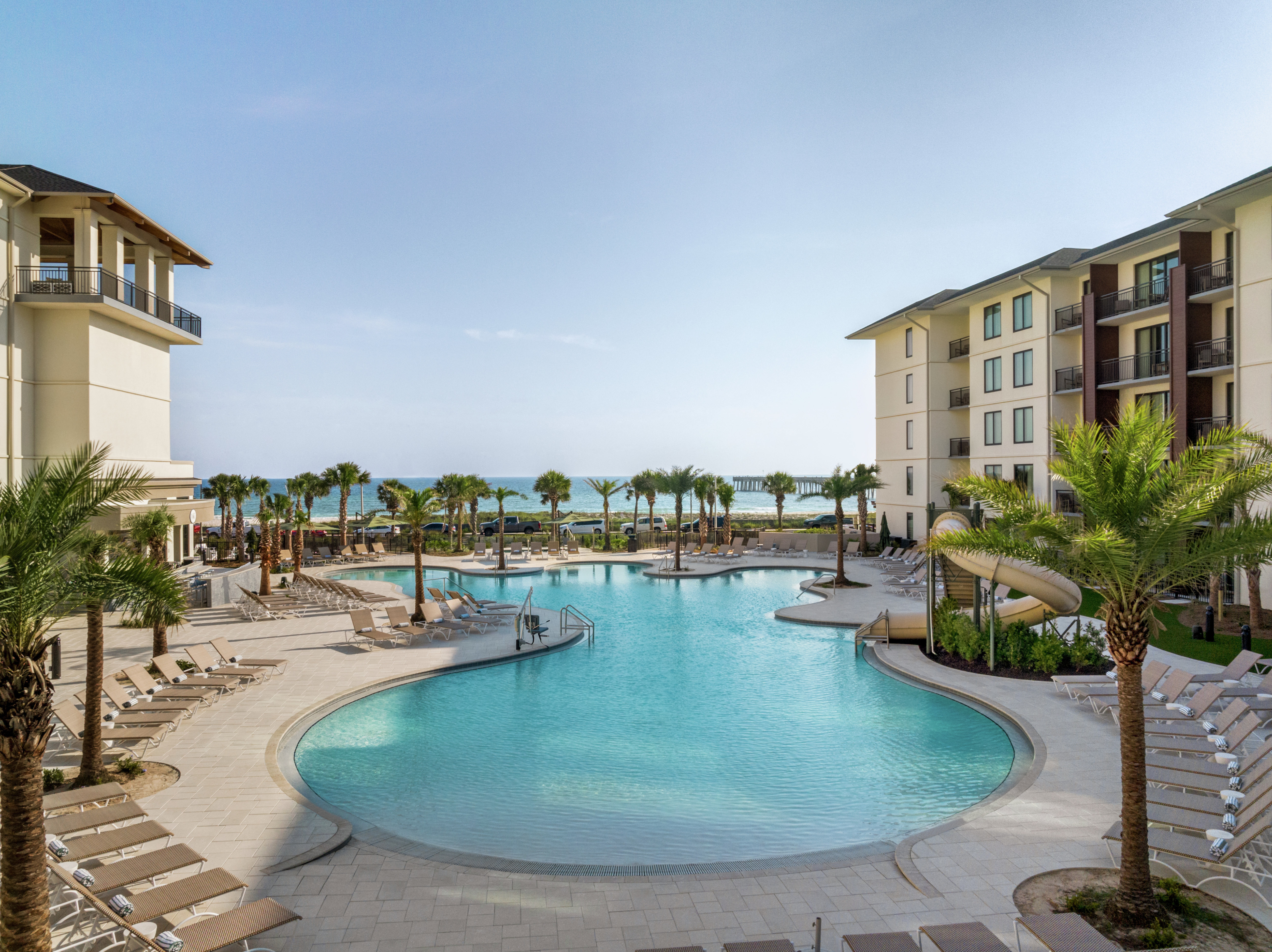 Beautiful resort pool deck with ample lounge seating, waterslide, lush palm trees and ocean views