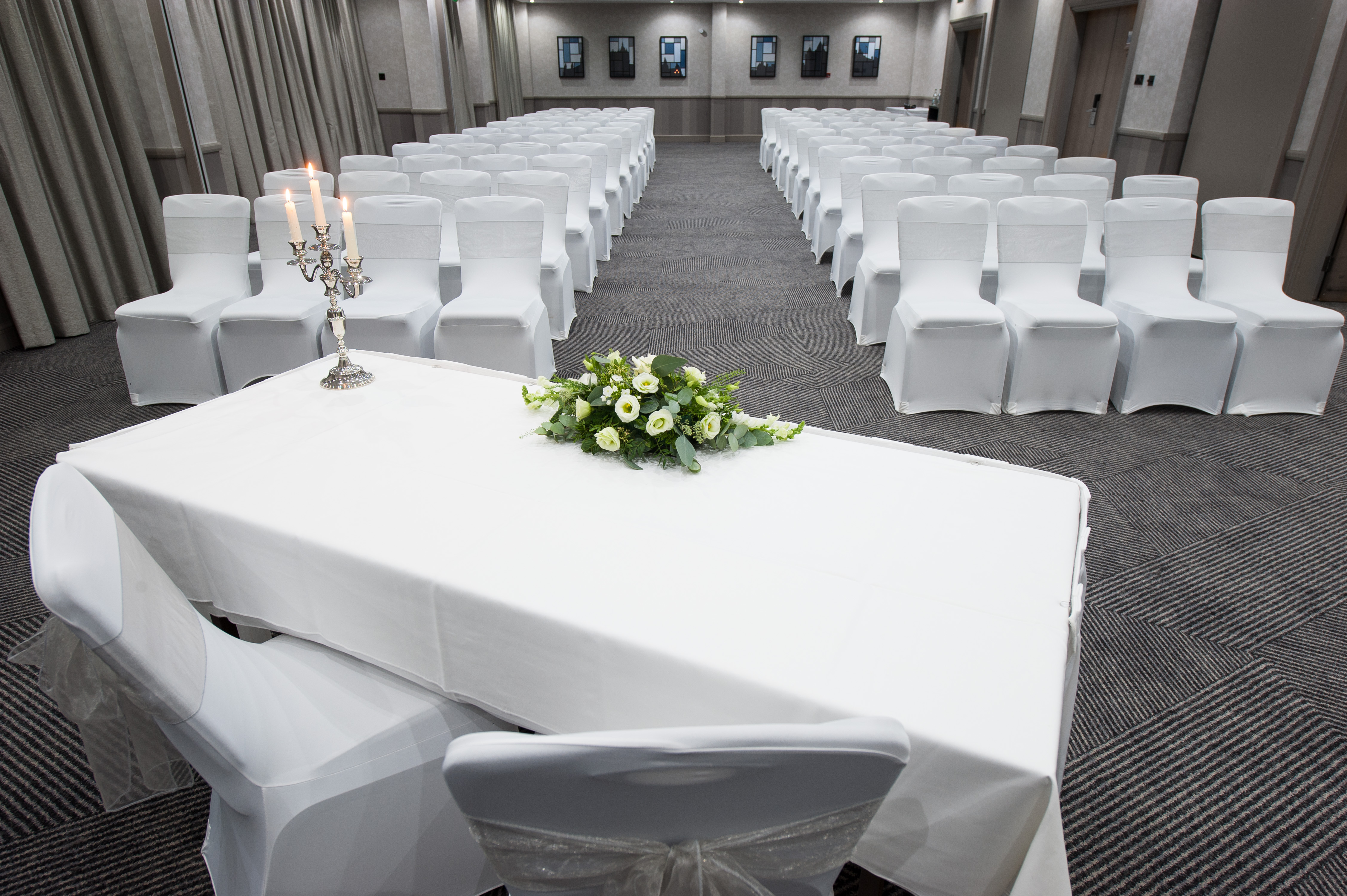 Front View of Meeting Room Arranged Theater Style With Rows of White Chairs Facing Table With Two Chairs, Candles and Flowers on White Linens