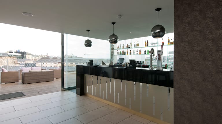 Decorative Lighting Above Service Area of Fully Stocked Bar, Soft Seating and Tables on Terrace With City View