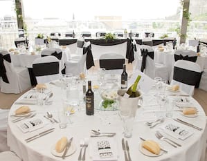 Overview of SkyBar Set Up for Wedding Reception With White Chairs With Black Bows, Tables With Place Settings, Bread, and Wine on White Linens, and Large Window With City View