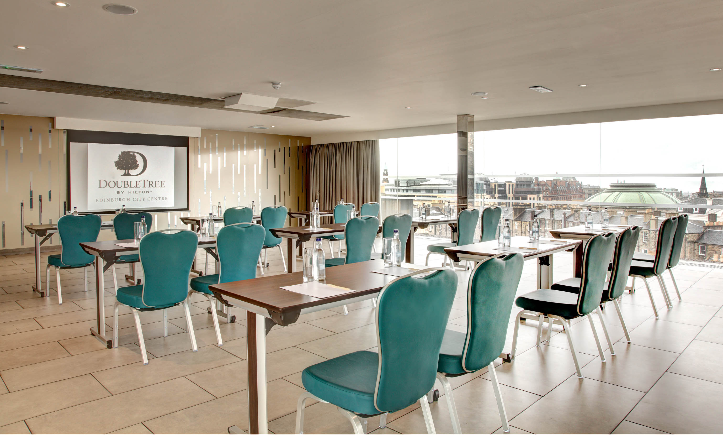 Classroom Setup in Penthouse Meeting Room With Tables and Green Chairs Facing Presentation Screen, Overhead Projector, and Large Window With Open Drapes to City View