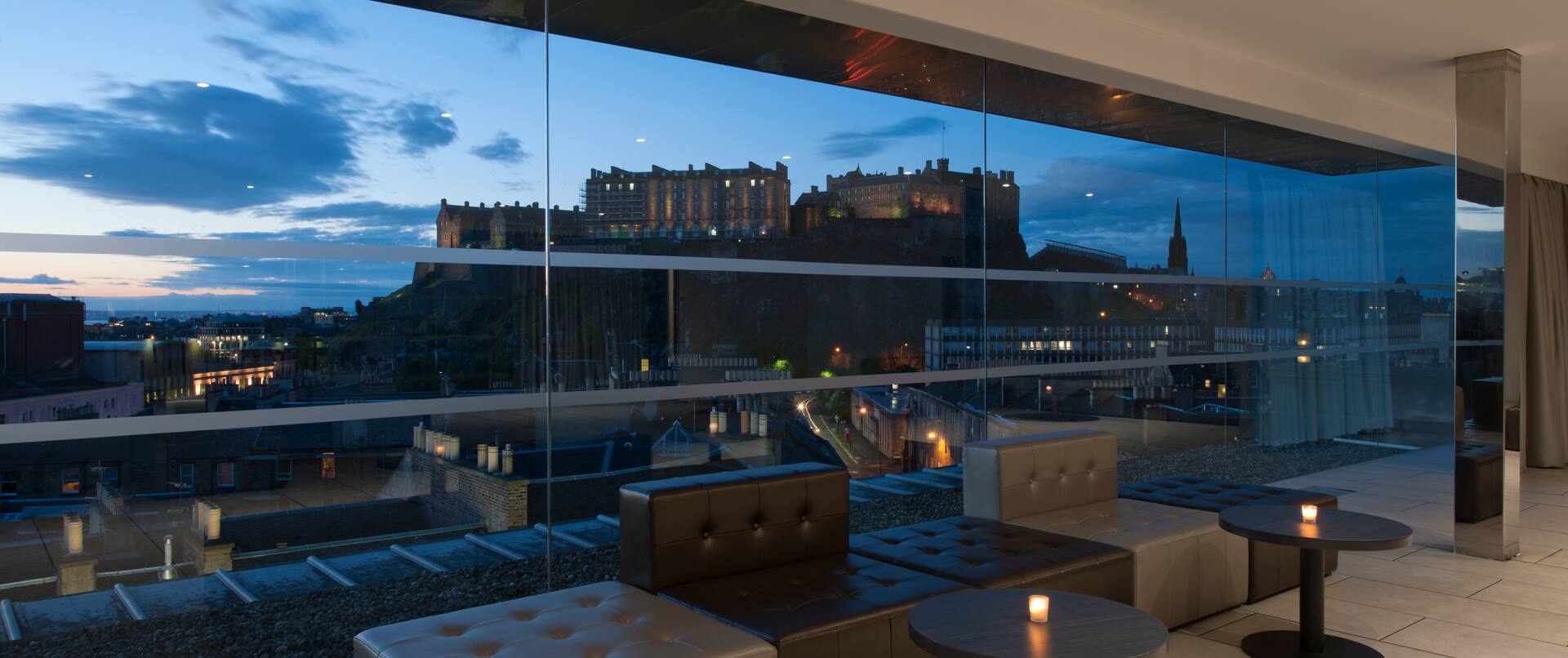 Tables With Illuminated Candles, and Lounge Seating by Large Window With View of Illuminated Castle at Dusk in SKYbar 