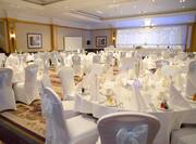 White Chairs With Bows, Round Tables With Place Settings and Flowers on White Linens, Presentation Screen and Large Window With Sheer Drapes in Strathallan Suite Set Up for Wedding Reception