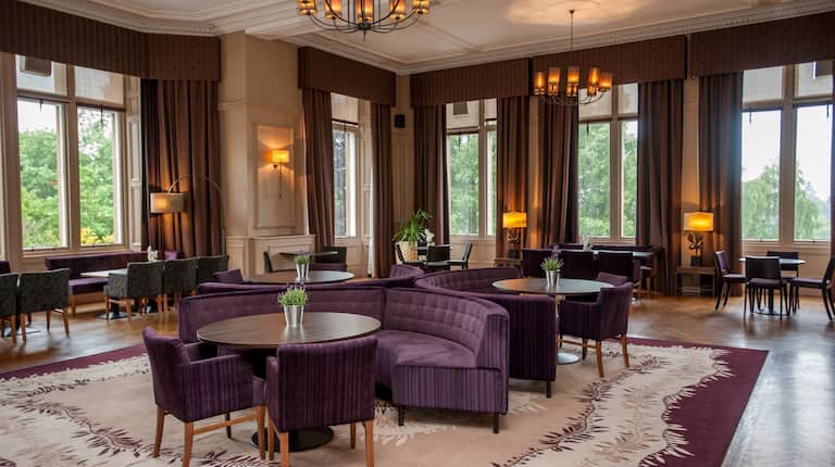 Tables. Semicircle Sofas, Armchairs, Illuminated Lamps, and Large Windows With Open Drapes in Stuart Lounge