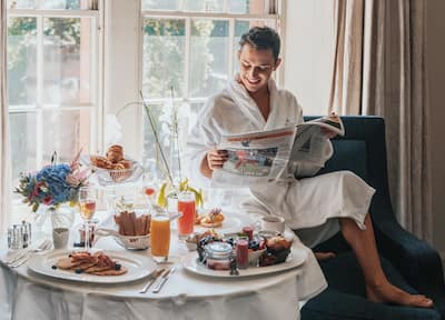 Man reading a newspaper by a large window and a hearty breakfast laid out in front of him