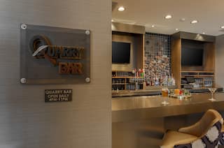 Detailed View of Quarry Bar and Hours of Operation Signage With View of TVs and Seating at Counter With Cocktails and Fully Stocked Bar