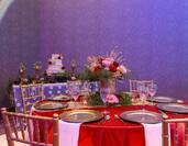 Chairs, Tables With Place Settings, White Napkins, and Flowers on Red Linens and Table With Candles and Wedding Cake in Ballroom Set Up for Wedding Reception