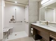 Spacious accessible bathroom featuring large vanity, mirror, roll-in shower with seat and grab bars.