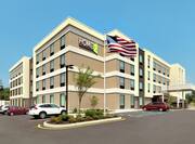 Modern Home2 Suites hotel exterior featuring ample parking, American flag flying proudly, and bright blue sky.