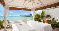Two massage tables set up under a shelter with azure sea and a jetty in the background.