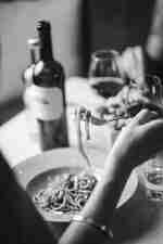 Mână care ține spaghete cu furculiță, cu sticlă de vin și pahare în fundal.