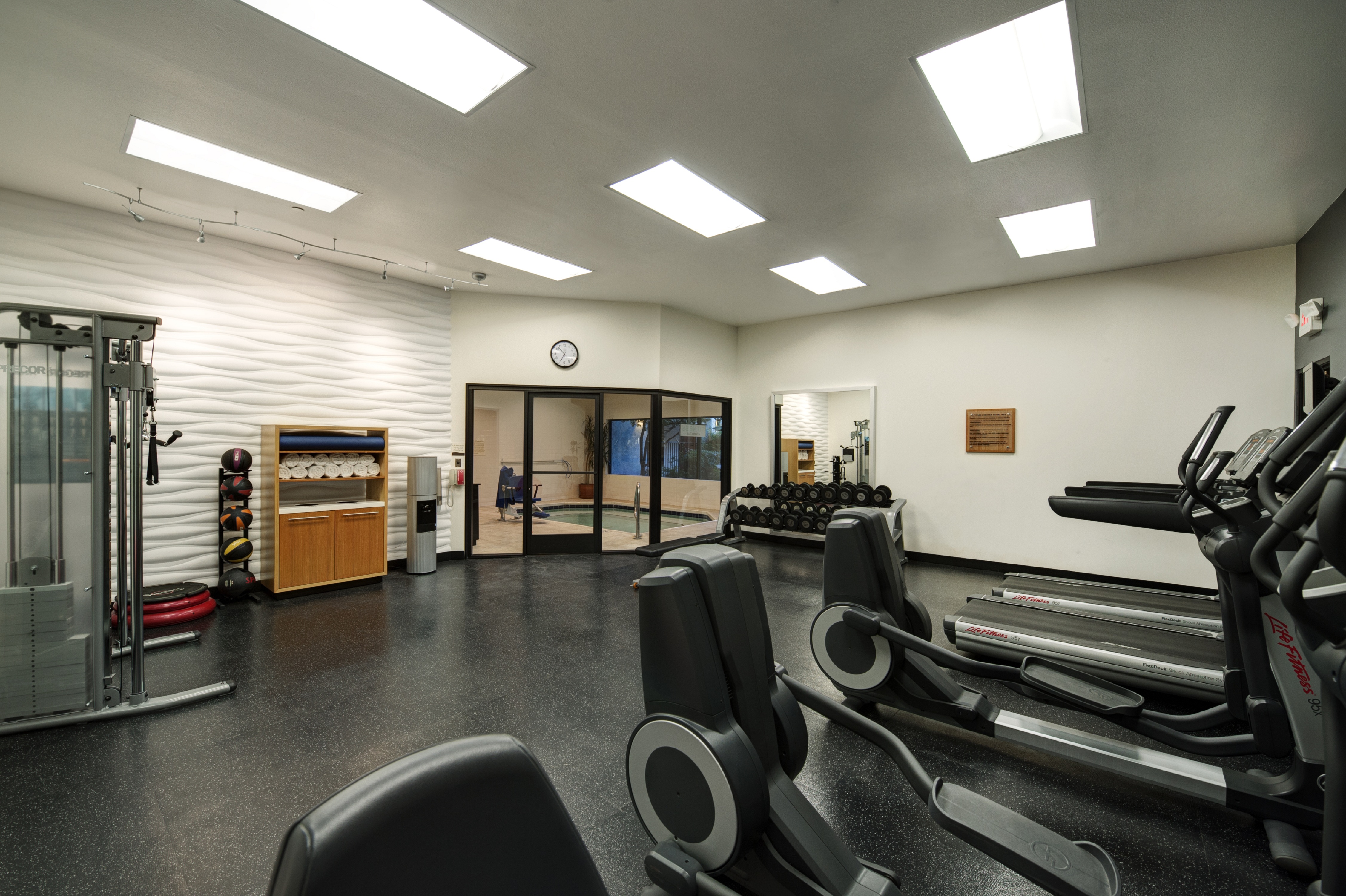 Fitness Center With Cardio Equipment, Weight Machine, Exercise Stepper, Weight Balls, Towel Station, Water Cooler, Wall Clock Above Glass Door With View of Hot Tub, and Large Mirror by Free Weight