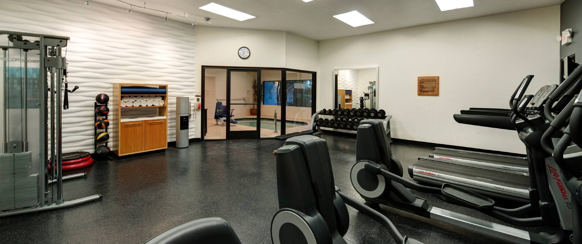 Fitness Center With Cardio Equipment, Weight Machine, Exercise Stepper, Weight Balls, Towel Station, Water Cooler, Wall Clock Above Glass Door With View of Hot Tub, and Large Mirror by Free Weight
