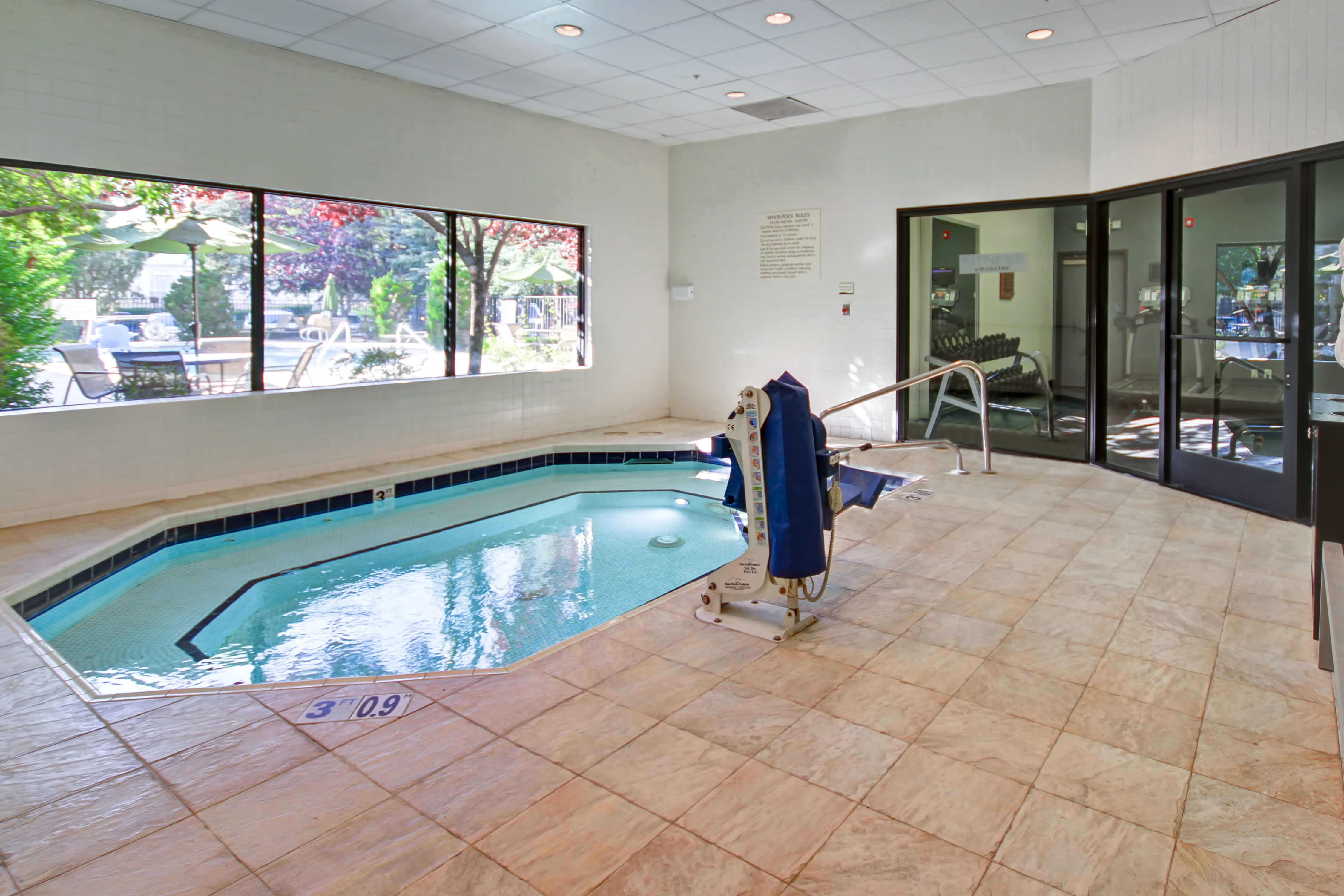 Indoor Whirlpool Spa By Large Window With Patio View and Glass Door With View Into Fitness Center