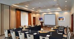 Meeting Room With Water Pitchers, Napkins, and Drinkware on U-Shaped Table With Black Linens, Chairs, Windows With Closed Drapes, Entry Doors, Wall Art, Podium and Presentation Screen