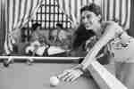 Woman playing pool in a bathing suit with a man and women sitting in a cabana behind her
