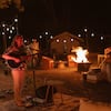 Mann mit Mikrofonständer spielt Gitarre und singt. Vier Männer und Frauen entspannen im Hintergrund vor einem Lagerfeuer und hören zu.