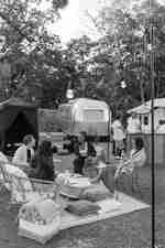 Three women and one man sitting around a coffee table outside laughing and talking while two men order at an outdoor bar with a femaler bartender. The setting is a campground in  a forest with two luxury tents and one airstream.
