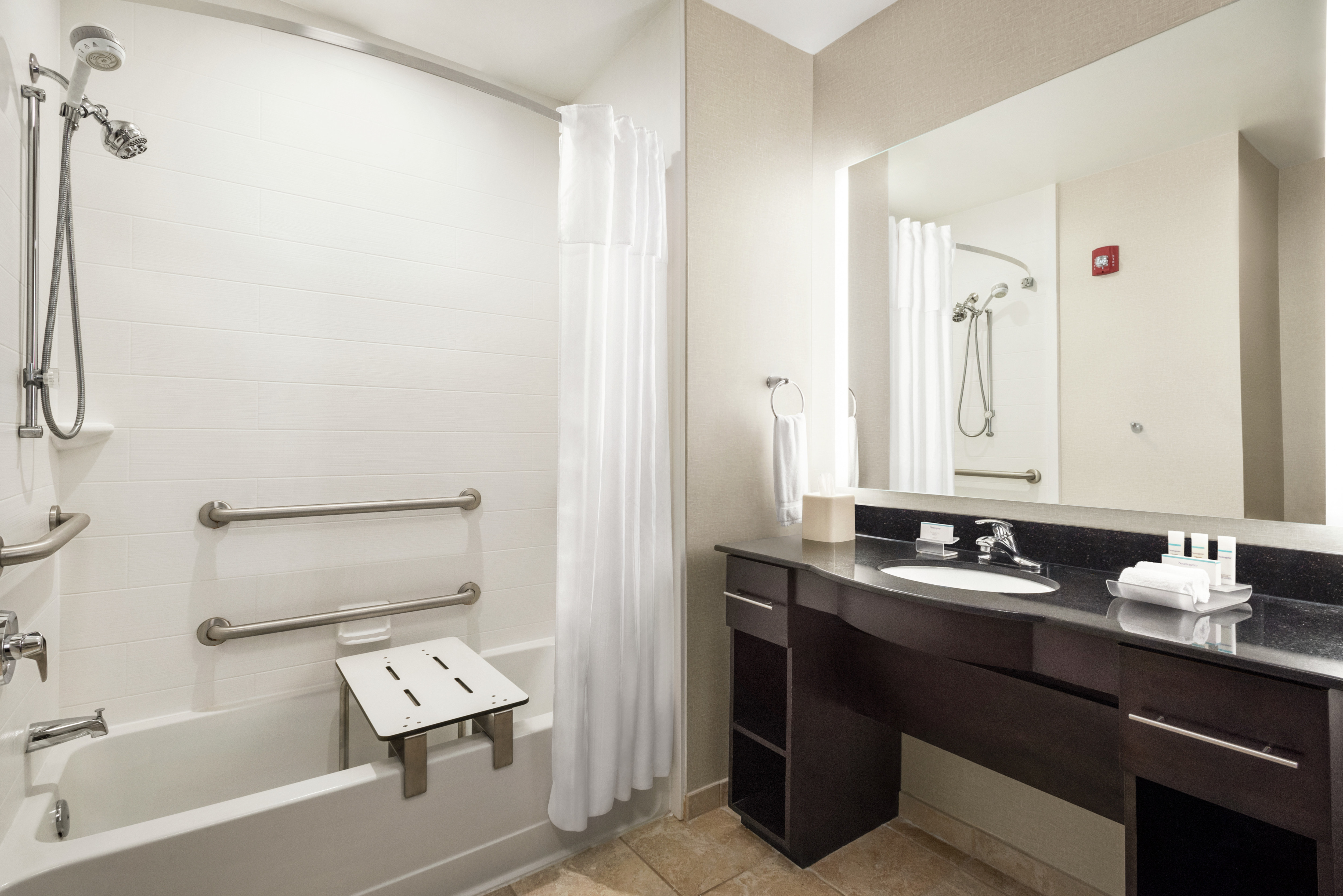 Spacious accessible bathroom featuring accessible tub with chair, large vanity and mirror with lights.