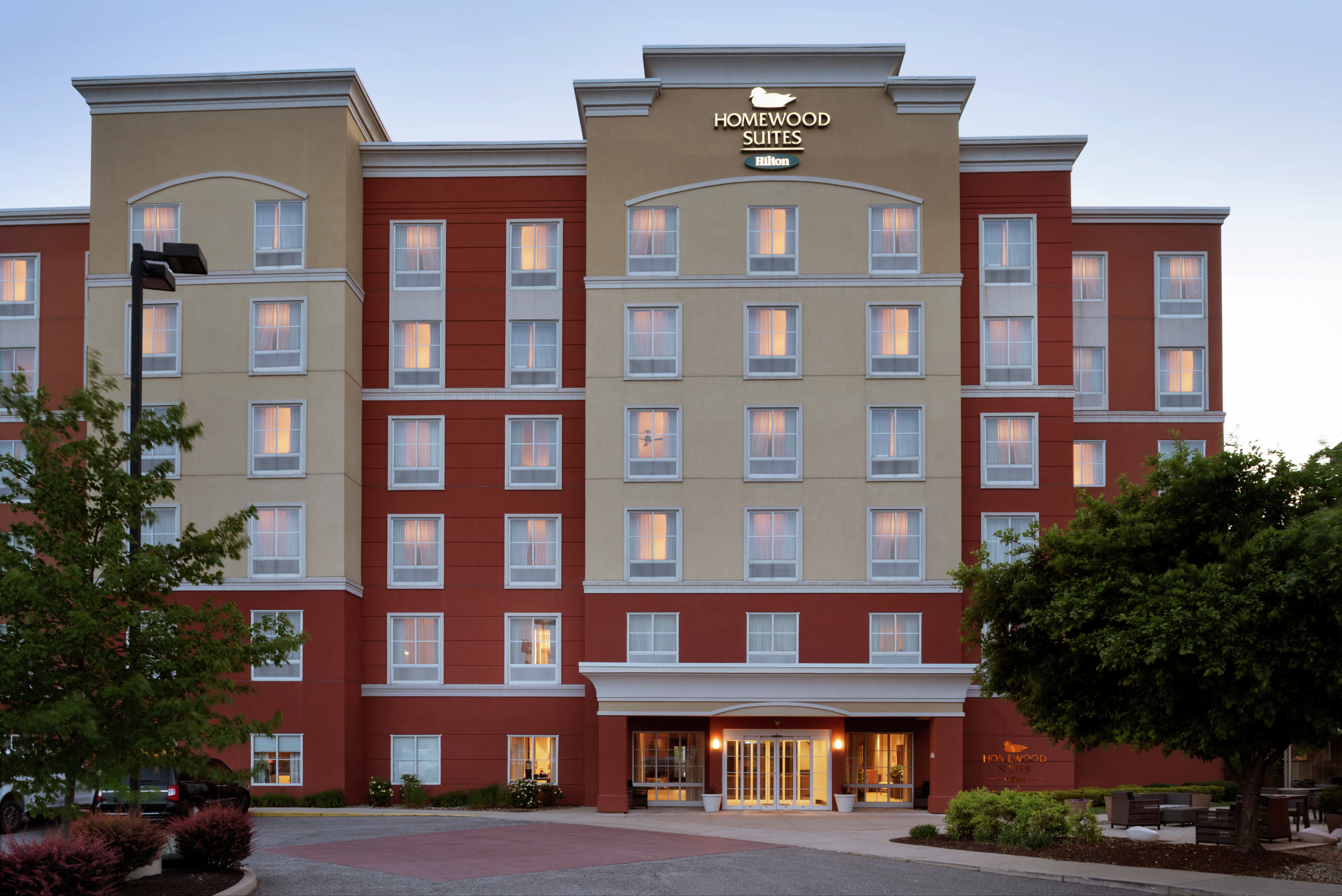 Welcoming Homewood Suites hotel exterior featuring glowing guest rooms, beautiful dusk sky, and lush trees.