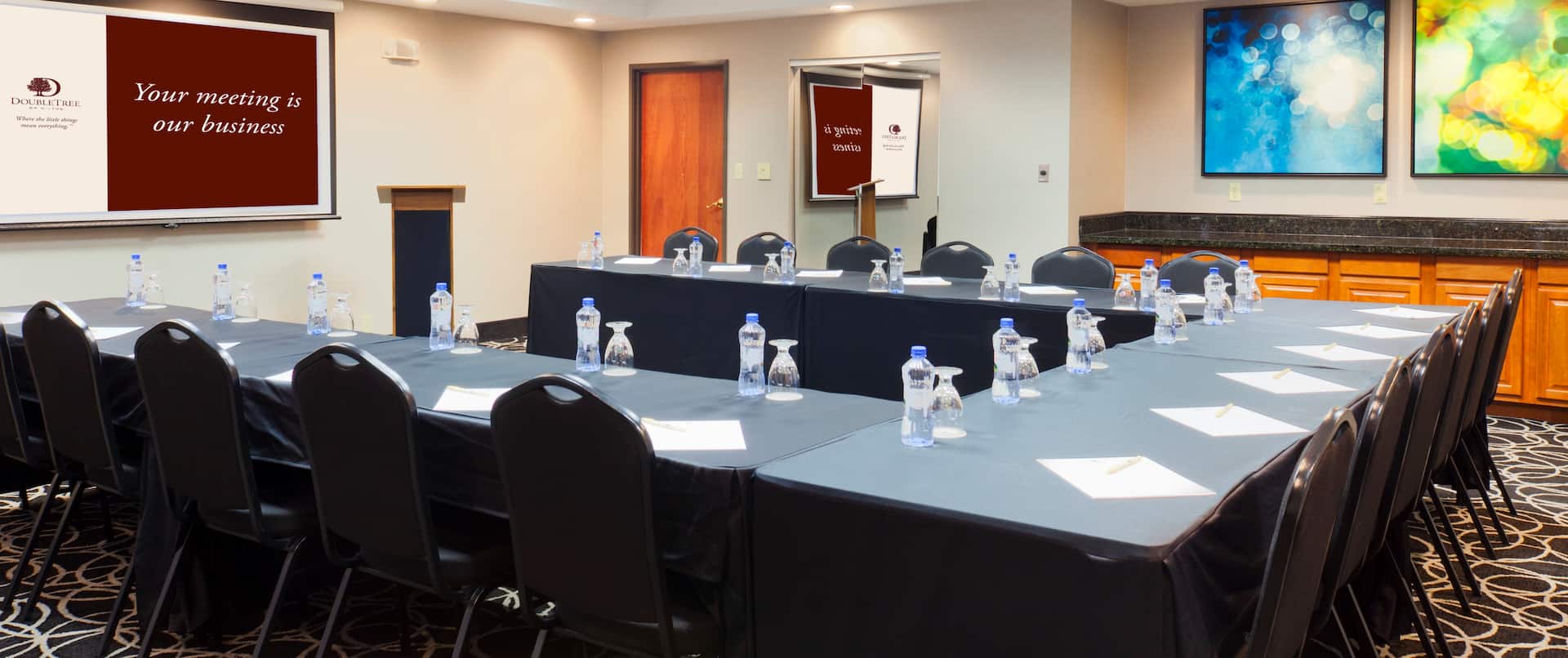 Meeting Room With Water Bottles and Drinking Glasses on U-Shaped Table, Chairs, TV, Podium, Entry, Wall Mirror, and Wall Art