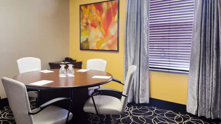 Wall Art, Window With Closed Blinds, Seating for Four at Round Table With Notepads and Drinking Glasses in  Small Conference Room