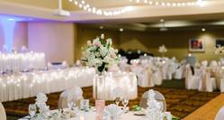  Place Settings, Candles, Flowers, Fan-Folded Napkins, White Linens on Dining Tables, and White Chairs With View of Dance Floor and Head Table in Background of Meeting Room Set Up for Wedding Reception