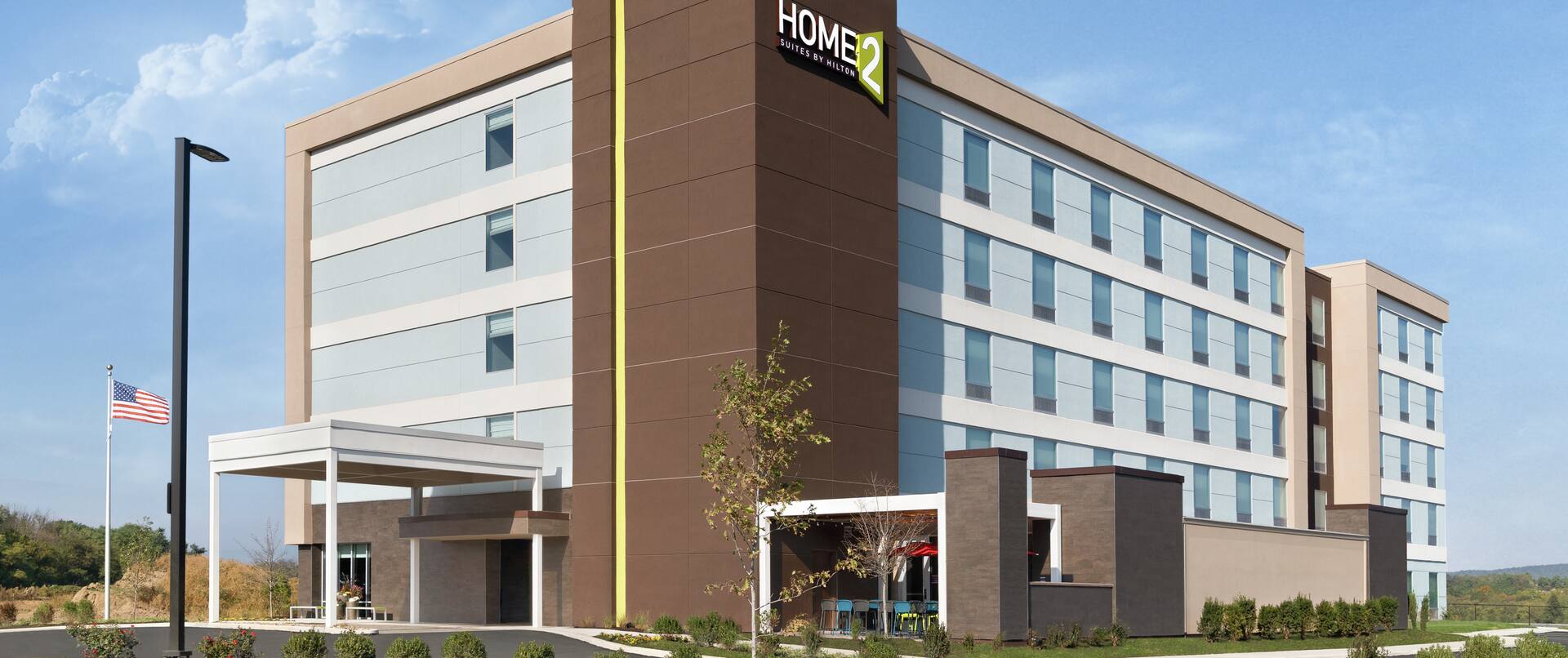 Modern Home2 Suites Hotel exterior featuring porte cochere, American flag on pole, and bright blue sky.
