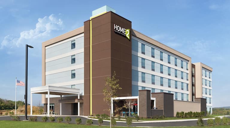 Modern Home2 Suites Hotel exterior featuring porte cochere, American flag on pole, and bright blue sky.