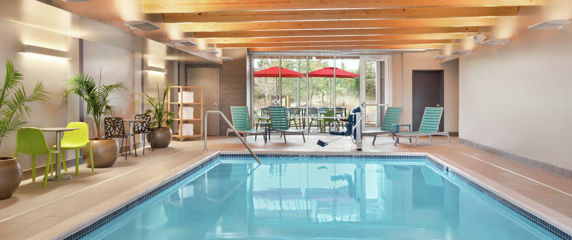 Indoor swimming pool featuring lush plants, lounge chairs, accessible chair lift, and beautiful outside view.