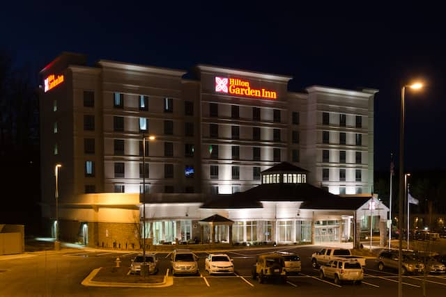 Hotel Exterior, Signage, Circle Driveway, Flag Pole, Landscaping, and Guest Cars on Parking Lot Illuminated at Night