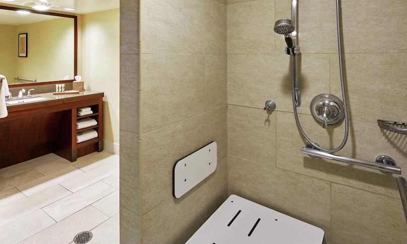 Salle de bain d'une chambre Diamond Head pour personnes à mobilité réduite avec miroir, comptoir lavabo et douche accessible en fauteuil roulant avec siège, transition précédente