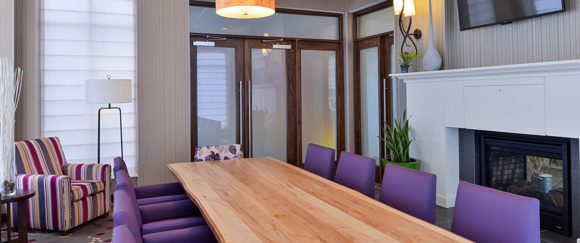 Conference Room in Boardroom Setup With TV, Fireplace, Long Table Surrounded by Purple Chairs, and Arm Chair in Corner by Windows 