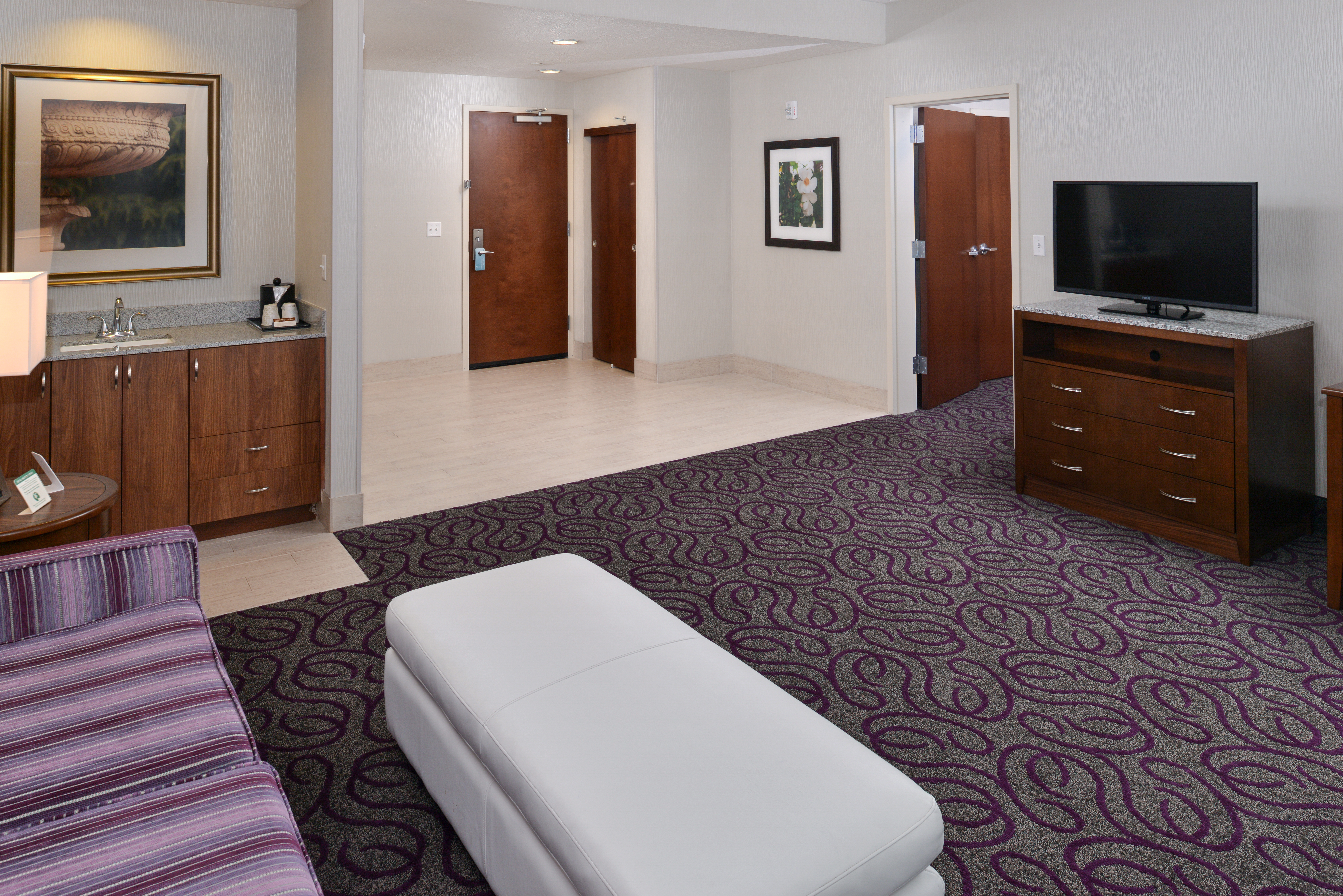 Living Suite With Wet Bar Area, Entry, Wall Art, Open Doorway to Bedroom, TV, Sofa, and Ottoman