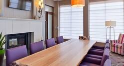 Conference Room with TV Above Fireplace, Large Windows, Long Table, Ten Purple Chairs and Armchair in Corner  by Window
