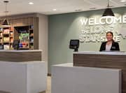 Hotel lobby front desk featuring welcoming staff member, Hilton Honors sign, and convenient treat shop.
