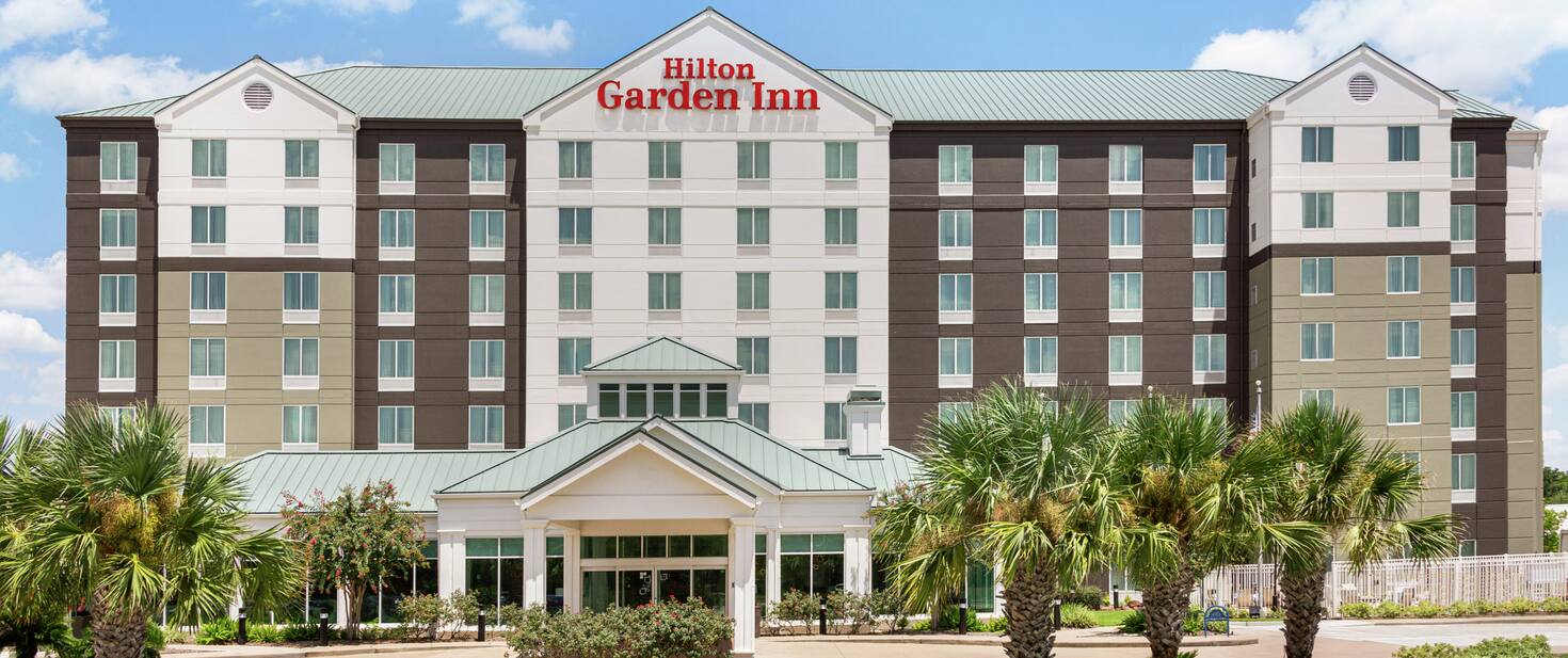 Welcoming Hilton Garden Inn hotel exterior featuring porte cochere, lush palm trees, and bright blue sky.