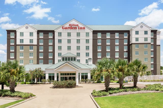 Welcoming Hilton Garden Inn hotel exterior featuring porte cochere, lush palm trees, and bright blue sky.