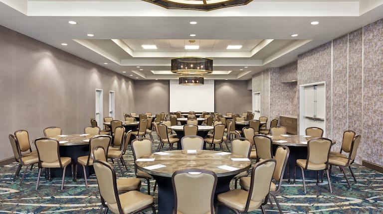 Spacious on-site ballroom fully set with banquet round tables and project screen at front of room.