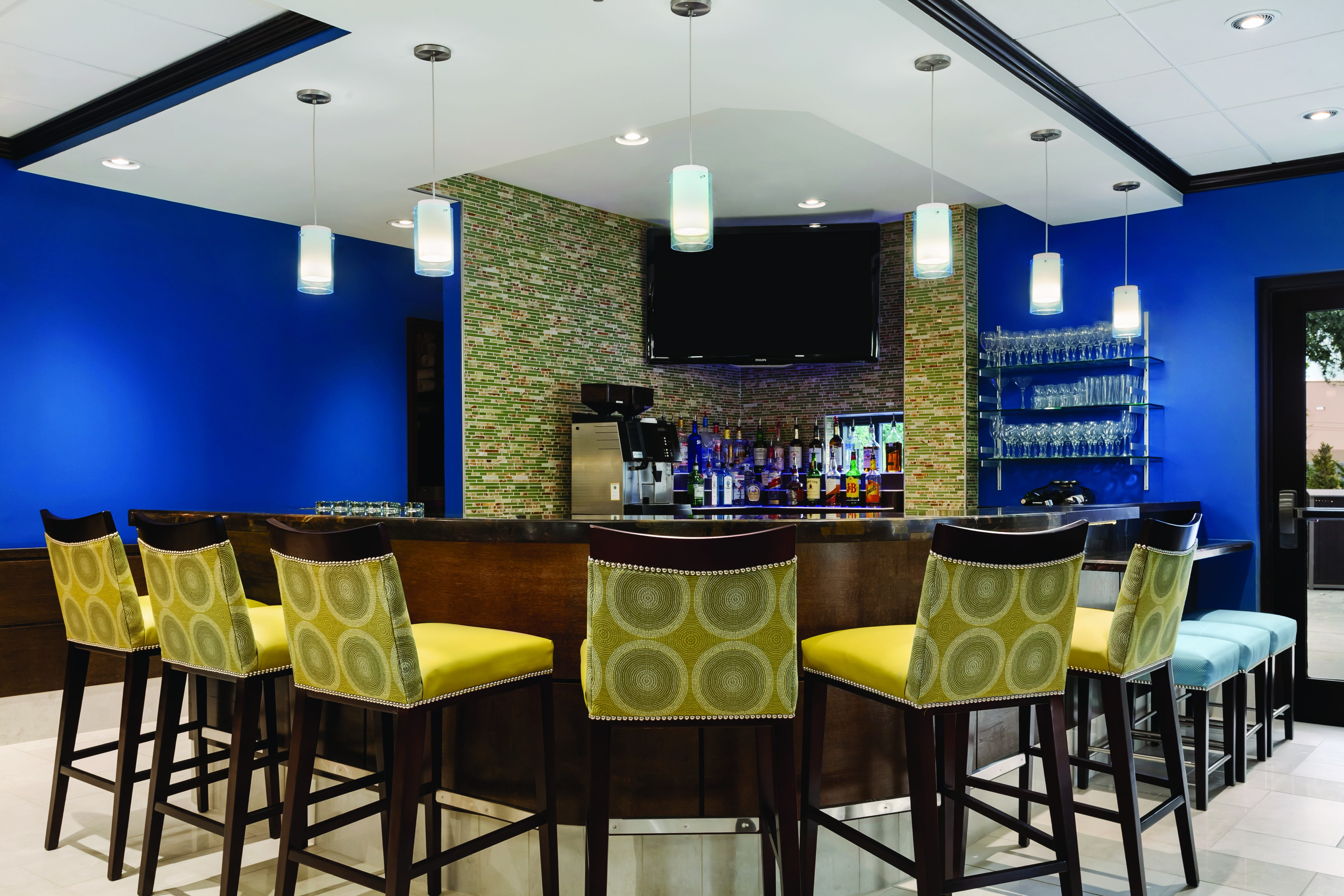 TV Above Fully Stocked Bar With Six Yellow Bar Chairs, and Three Blue Stools