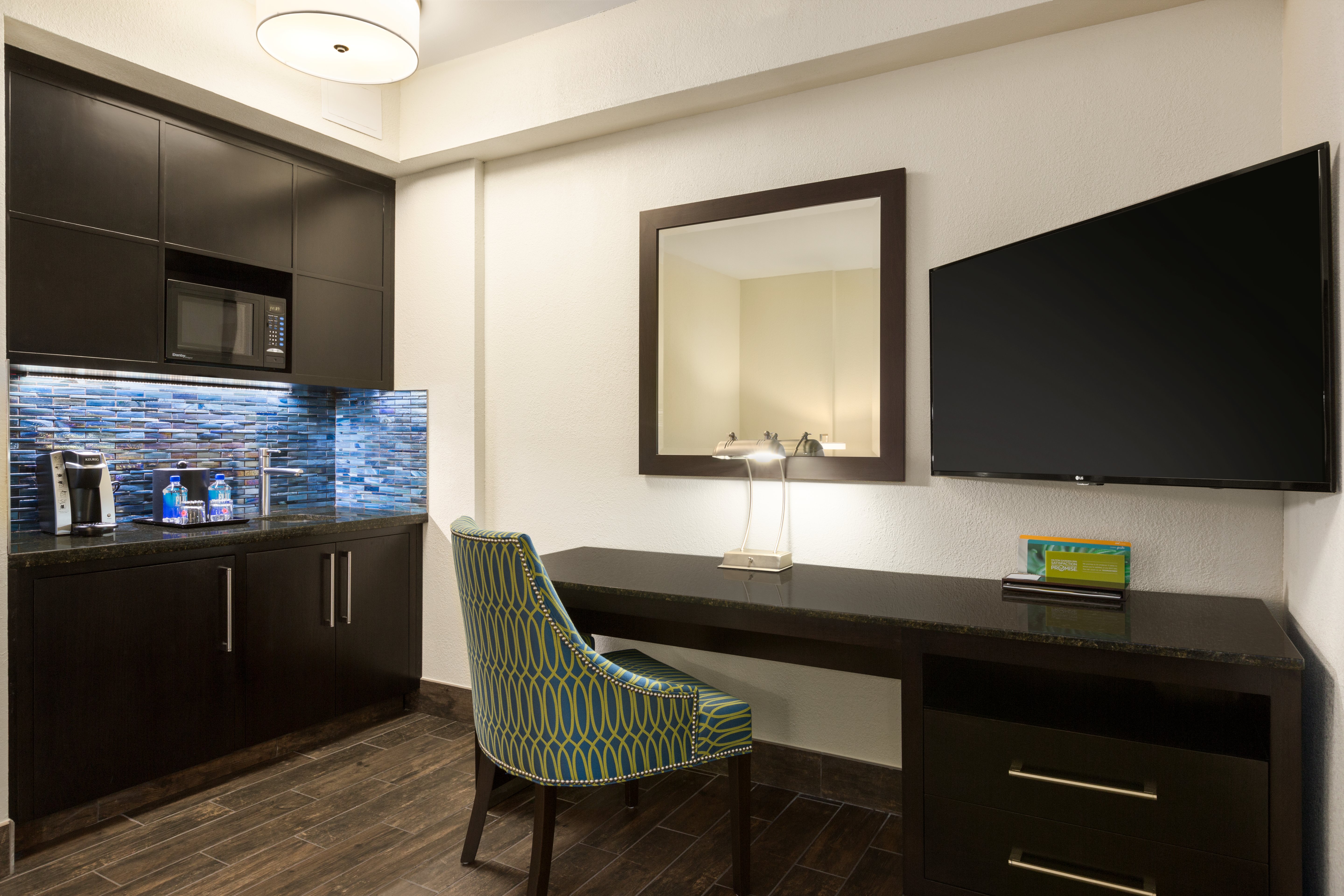 Wet Bar Area With Keurig, Bottled Water, Ice Bucket, Microwave, and Sink and TV Above Work Desk in Corner.of Junior Suite