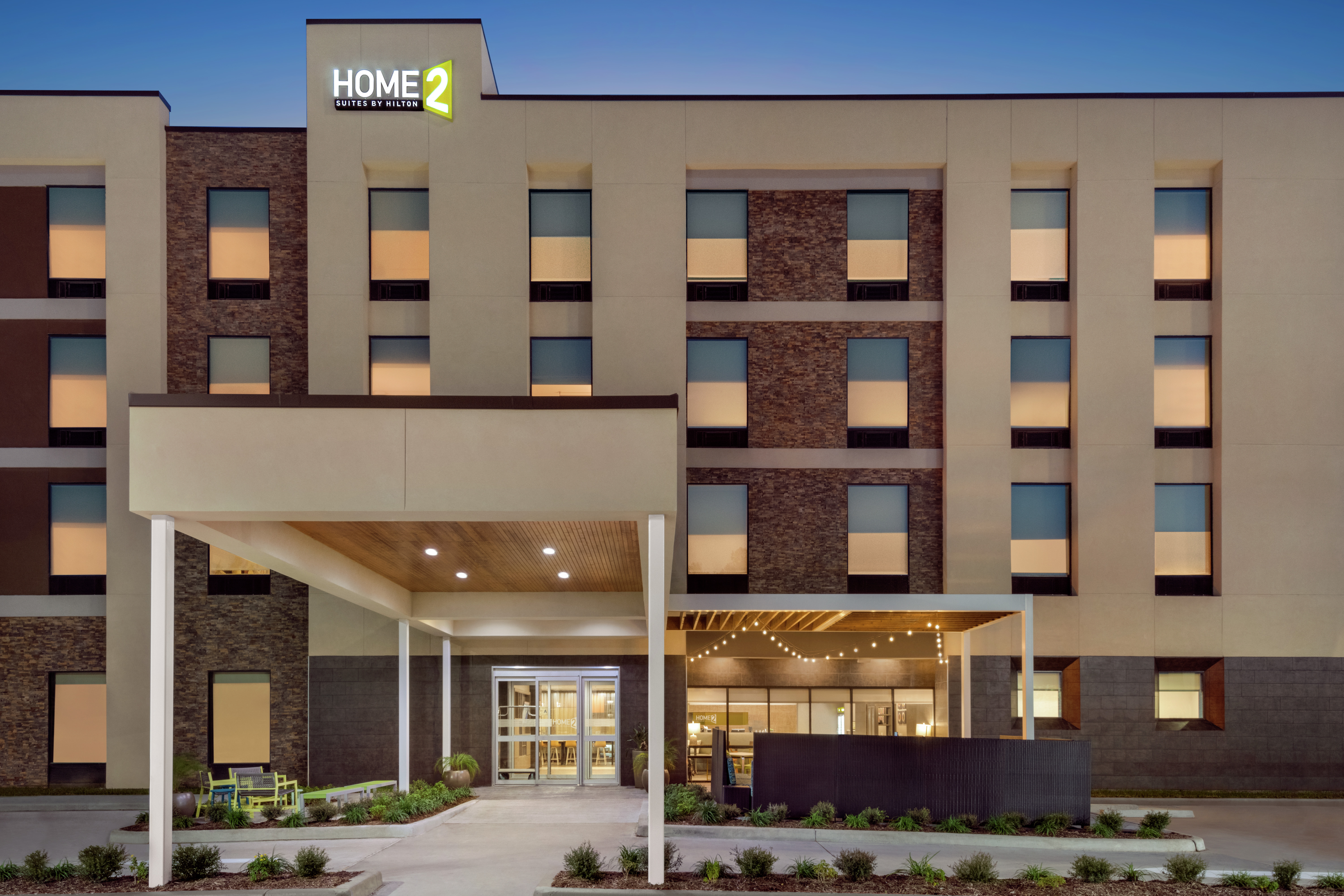Modern Home2 Suites hotel exterior with porte cochere, patio for guests to relax, and glowing lobby.
