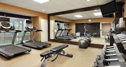 Bright fitness center featuring floor to ceiling mirror, cardio machines, free weights, TV, and complimentary towels. 