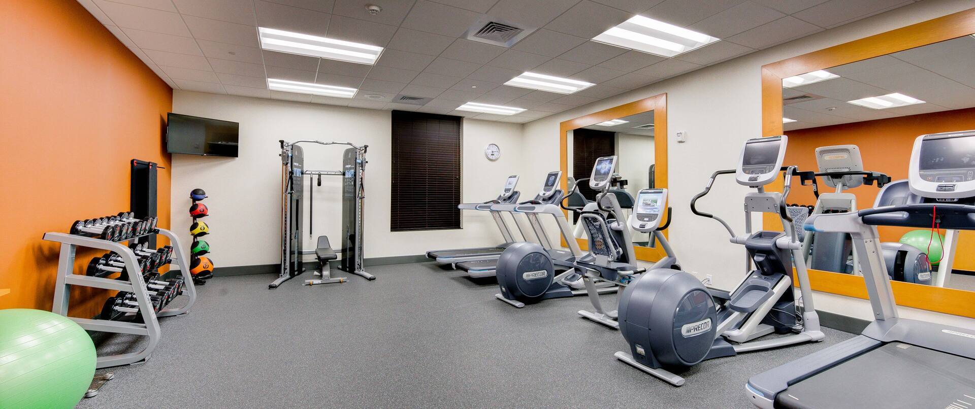 Fitness Center With Green Exercise Ball, Free Weights, TV Above Weight Balls, Weight Machine With Bench, and Cardio Equipment Facing Two Large Mirrors